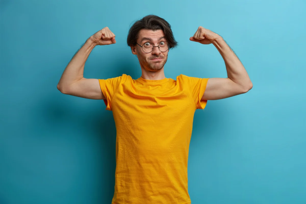 self confident powerful man raises arms shows muscles demonstrates result regular workout dressed yellow t shirt spectacles leads active healthy lifestyle being very strong