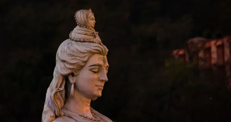 A close-up statue of Lord Shiva with a smaller figure atop his head, illuminated against a dark background.