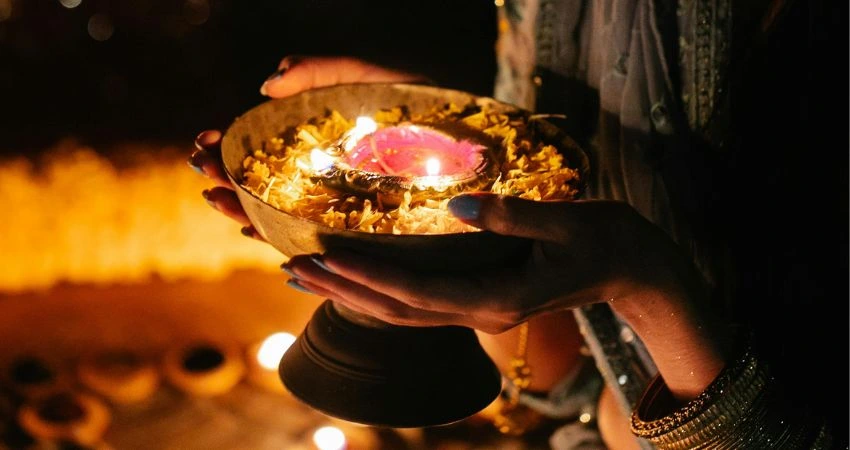 Hands holding a lit diya (oil lamp) in a decorative bowl, surrounded by flower petals, in a dark setting.